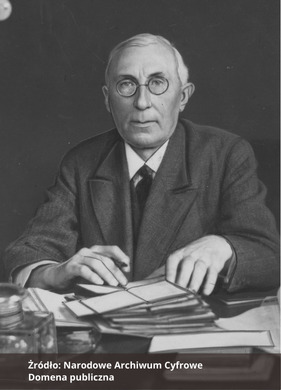 Fotografia czarno-biała przedstawiająca portret mężczyzny z okularami, siedzącego przy biurku, ubranego elegancko. Na biurku leżą dokumenty. Na dole zdjęcia napis "Źródło: Narodowe Archiwum Cyfrowe oraz Domena publiczna".