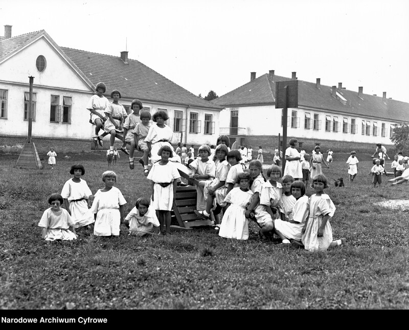 Czarno-biała fotografia pochodzi ze zbiorów Narodowego Archiwum Cyfrowego, ukazuje dzieci szkolne z lat 1920–1930. Zdjęcie wykonano na szerokim, trawiastym placu przed długim budynkiem w stylu szkolnym z wieloma oknami i dwuspadowym dachem. Budynek przypomina dawną szkołę lub placówkę edukacyjną. W centrum znajduje się kilkanaścioro dzieci (głównie dziewczynek), ubranych w jasne, proste sukienki z krótkimi rękawami. Fryzury dzieci są jednolite: krótkie, równo ścięte włosy. Część dzieci stoi, część siedzi albo klęczy na trawie, kilkoro bawi się na drewnianej huśtawce. Po prawej dzieci są ujęte w ruchu na huśtawce lub trampolinie, jakby w trakcie skoku. Po prawej kilkoro dzieci siedzi obok drewnianego urządzenia zabawowego. Na dalszym planie widać inne dzieci spacerujące, być może pod opieką dorosłych. Scena oddaje atmosferę wspólnotowej, radosnej zabawy, typową dla zorganizowanych zajęć lub kolonii letnich.