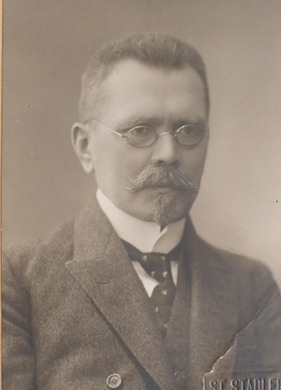 Ilustracja czarno-biała przedstawiająca mężczyznę w okularach z wąsami i brodą, ubranego elegancko, uchwyconego z prawnego profilu. 