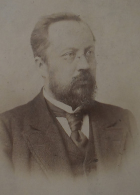 Fotografia w sepii popiersia mężczyzny w okularach z brodą i wąsami, elegancko ubranego, przedstawionego z prawego profilu. 