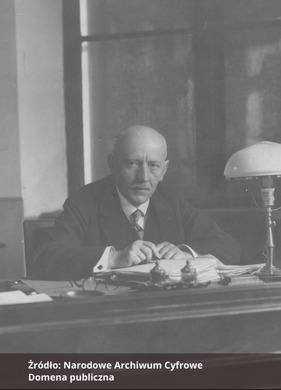 Fotografia czarno-biała przedstawiająca portret mężczyzny z wąsami, ubranego elegancko i siedzącego przy biurku. Na dole zdjęcia napis "Źródło: Narodowe Archiwum Cyfrowe oraz Domena publiczna".
