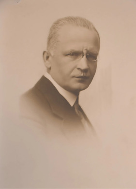 Fotografia w sepii przedstawiająca mężczyznę w okularach z wąsami, elegancko ubranego, przedstawionego z prawego profilu.