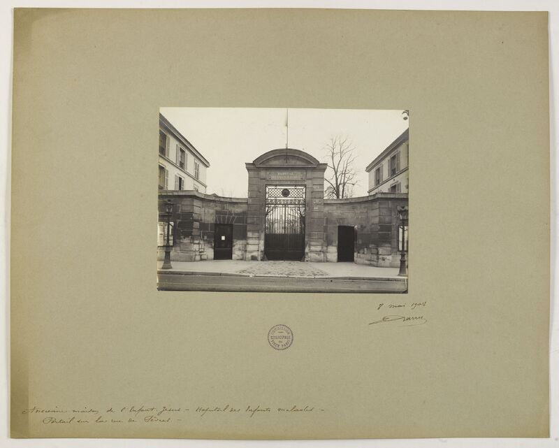 Zdjęcie przedstawia czarno-białą fotografię historyczną.
Przedstawia główną bramę obiektu – szpitala dziecięcego w Paryżu, otoczoną ceglanym murem i symetrycznie usytuowanymi bocznymi budynkami. Brama ma łukowaty zwieńczony portal z napisem „HÔPITAL DES ENFANTS MALADES” tuż nad kratą wejściową. Obiekt stoi przy ulicy, widoczny jest fragment chodnika i jezdni.
Po obu stronach portalu rozciągają się niskie ściany, w które wbudowane są dwa mniejsze wejścia – po jednym z lewej i prawej strony głównej bramy. Całość ma klasycystyczny charakter. Za bramą widać gołe drzewa i fragment budynku. Pomiędzy bramą a fotografującym jest otwarta przestrzeń – prawdopodobnie plac przed wejściem. Na kartonie poniżej widać odręczny podpis datowany na 8 maja 1908 oraz pieczątkę archiwalną.