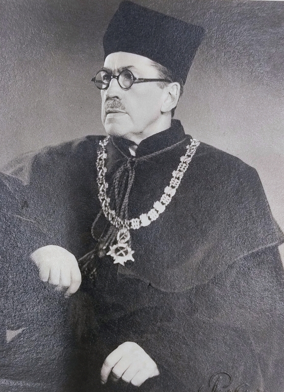 Zdjęcie przedstawia dorosłego mężczyznę z głową i wzrokiem zwróconymi w lewo. Na twarzy ma wąsy oraz okrągłe okulary. Ubrany jest w ciemną szatę z wysokim kołnierzem oraz charakterystyczne nakrycie głowy. Na szyi zawieszony jest ozdobny łańcuch z dużych ogniw, zakończony medalionem. Prawa ręka jest lekko uniesiona w górę, lewa spoczywa trochę niżej.