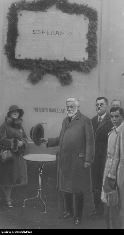 Grupa ludzi stoi wokół stołu, w centrum starszy mężczyzna w płaszczu, trzymający kapelusz w dłoni, powyżej tablica z napisem esperanto.