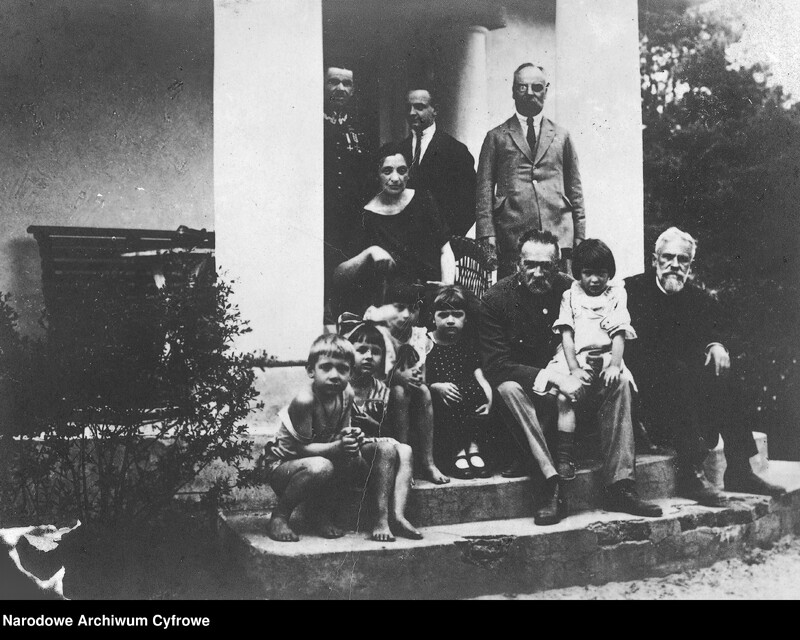 Grupa osób przebywająca na schodach prowadzących do wejścia do dworku. W części centralnej siedzi mężczyzna trzymający na kolanach dziecko, obok grupka dzieci oraz starszy mężczyzna z brodą. Powyżej kobieta z laską siedząca w wiklinowym fotelu oraz trzech stojących mężczyzn, jeden z nich ubrany w mundur wojska polskiego.