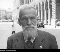 Starszy mężczyzna z brodą , ubrany w garnitur stoi przed budynkiem z arkadami.