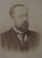 Fotografia w sepii popiersia mężczyzny w okularach z brodą i wąsami, elegancko ubranego, przedstawionego z prawego profilu. 