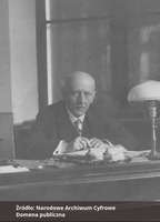 Fotografia czarno-biała przedstawiająca portret mężczyzny z wąsami, ubranego  elegancko i siedzącego przy biurku. Na dole zdjęcia napis "Źródło: Narodowe Archiwum Cyfrowe oraz Domena publiczna".