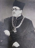 Zdjęcie przedstawia dorosłego mężczyznę z głową i wzrokiem zwróconymi w lewo. Na twarzy ma wąsy oraz okrągłe okulary. Ubrany jest w ciemną szatę z wysokim kołnierzem oraz charakterystyczne nakrycie głowy. Na szyi zawieszony jest ozdobny łańcuch z dużych ogniw, zakończony medalionem. Prawa ręka jest lekko uniesiona w górę, lewa spoczywa trochę niżej.