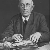 Fotografia czarno-biała przedstawiająca portret mężczyzny z okularami, siedzącego przy biurku, ubranego elegancko. Na biurku leżą dokumenty. Na dole zdjęcia napis "Źródło: Narodowe Archiwum Cyfrowe oraz Domena publiczna".