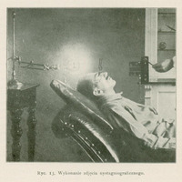 Zdjęcie przedstawia scenę związaną z wykonaniem zdjęcia nystagmograficznego, czyli badania ruchów gałek ocznych.
Widać na nim mężczyznę w średnim wieku, leżącego na odchylonym do tyłu fotelu, podobnym do fotela dentystycznego. Głowa pacjenta jest lekko odchylona do tyłu i unieruchomiona. Ma on na twarzy założone nasadki oczne, do których przymocowane są dwa cienkie pręciki (po jednym przed każdą gałką oczną).
Z lewej strony zdjęcia znajduje się lampa na wysokim statywie, skierowana bezpośrednio na oczy pacjenta — jej światło służy do tworzenia cieni pręcików na szczelinie światła lub filmie. Po prawej stronie widoczna jest ręka osoby (prawdopodobnie lekarza), która obsługuje urządzenie.
Tło zdjęcia stanowi wnętrze pomieszczenia — być może jest to gabinet okulistyczny lub laboratorium.