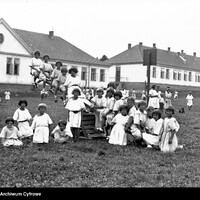 Czarno-biała fotografia pochodzi ze zbiorów Narodowego Archiwum Cyfrowego, ukazuje dzieci szkolne z lat 1920–1930. Zdjęcie wykonano na szerokim, trawiastym placu przed długim budynkiem w stylu szkolnym z wieloma oknami i dwuspadowym dachem. Budynek przypomina dawną szkołę lub placówkę edukacyjną. W centrum znajduje się kilkanaścioro dzieci (głównie dziewczynek), ubranych w jasne, proste sukienki z krótkimi rękawami. Fryzury dzieci są jednolite: krótkie, równo ścięte włosy. Część dzieci stoi, część siedzi albo klęczy na trawie, kilkoro bawi się na drewnianej huśtawce. Po prawej dzieci są ujęte w ruchu na huśtawce lub trampolinie, jakby w trakcie skoku. Po prawej kilkoro dzieci siedzi obok drewnianego urządzenia zabawowego. Na dalszym planie widać inne dzieci spacerujące, być może pod opieką dorosłych. Scena oddaje atmosferę wspólnotowej, radosnej zabawy, typową dla zorganizowanych zajęć lub kolonii letnich.