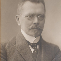 Ilustracja czarno-biała przedstawiająca mężczyznę w okularach z wąsami i brodą, ubranego elegancko, uchwyconego z prawnego profilu. 