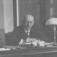 Fotografia czarno-biała przedstawiająca portret mężczyzny z wąsami, ubranego  elegancko i siedzącego przy biurku. Na dole zdjęcia napis "Źródło: Narodowe Archiwum Cyfrowe oraz Domena publiczna".