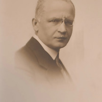 Fotografia w sepii przedstawiająca mężczyznę w okularach z wąsami, elegancko ubranego, przedstawionego z prawego profilu.