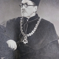 Zdjęcie przedstawia dorosłego mężczyznę z głową i wzrokiem zwróconymi w lewo. Na twarzy ma wąsy oraz okrągłe okulary. Ubrany jest w ciemną szatę z wysokim kołnierzem oraz charakterystyczne nakrycie głowy. Na szyi zawieszony jest ozdobny łańcuch z dużych ogniw, zakończony medalionem. Prawa ręka jest lekko uniesiona w górę, lewa spoczywa trochę niżej.