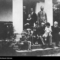 Grupa osób przebywająca na schodach prowadzących do wejścia do dworku. W części centralnej siedzi mężczyzna trzymający na kolanach dziecko, obok grupka dzieci oraz starszy mężczyzna z brodą. Powyżej kobieta z laską siedząca w wiklinowym fotelu oraz trzech stojących mężczyzn, jeden z nich ubrany w mundur wojska polskiego.