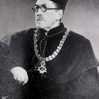 Fotografia czarno-biała przedstawiająca portret mężczyzny z wąsami, ubranego w togę oraz pelerynę i łańcuch. Na dole zdjęcia napis "Źródło: Muzeum Farmacji UJ CM oraz Domena publiczna".